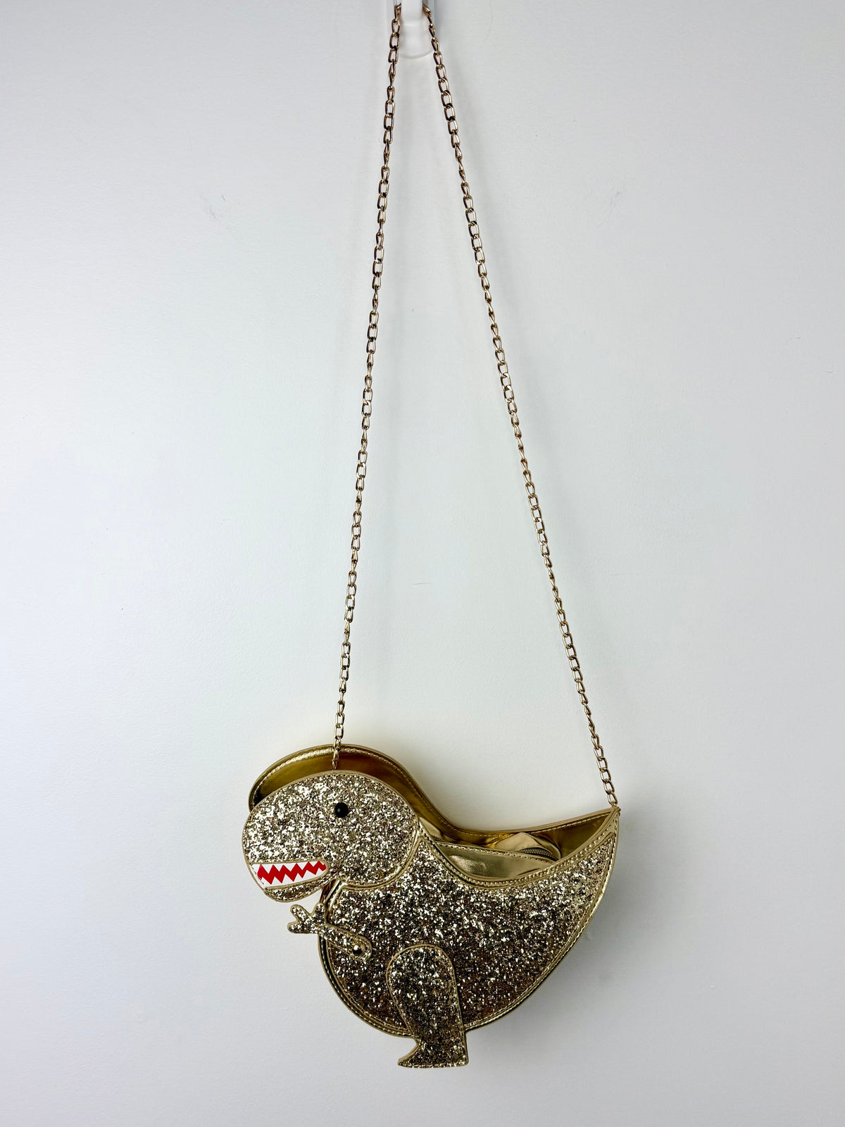 A gold glitter dinosaur-shaped clutch purse with a chain strap, featuring red and white enamel details on its teeth and a small black eye, shown against a plain white background.