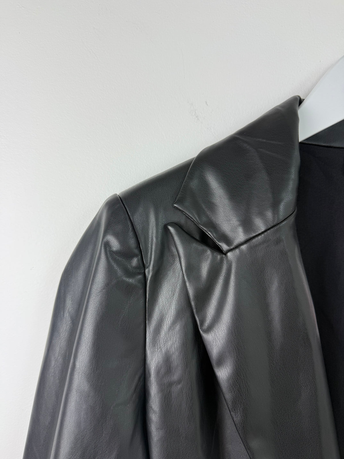 Close-up of a black faux-leather blazer jacket with a wide lapel and structured shoulder, hanging on a white hanger against a plain light gray wall.