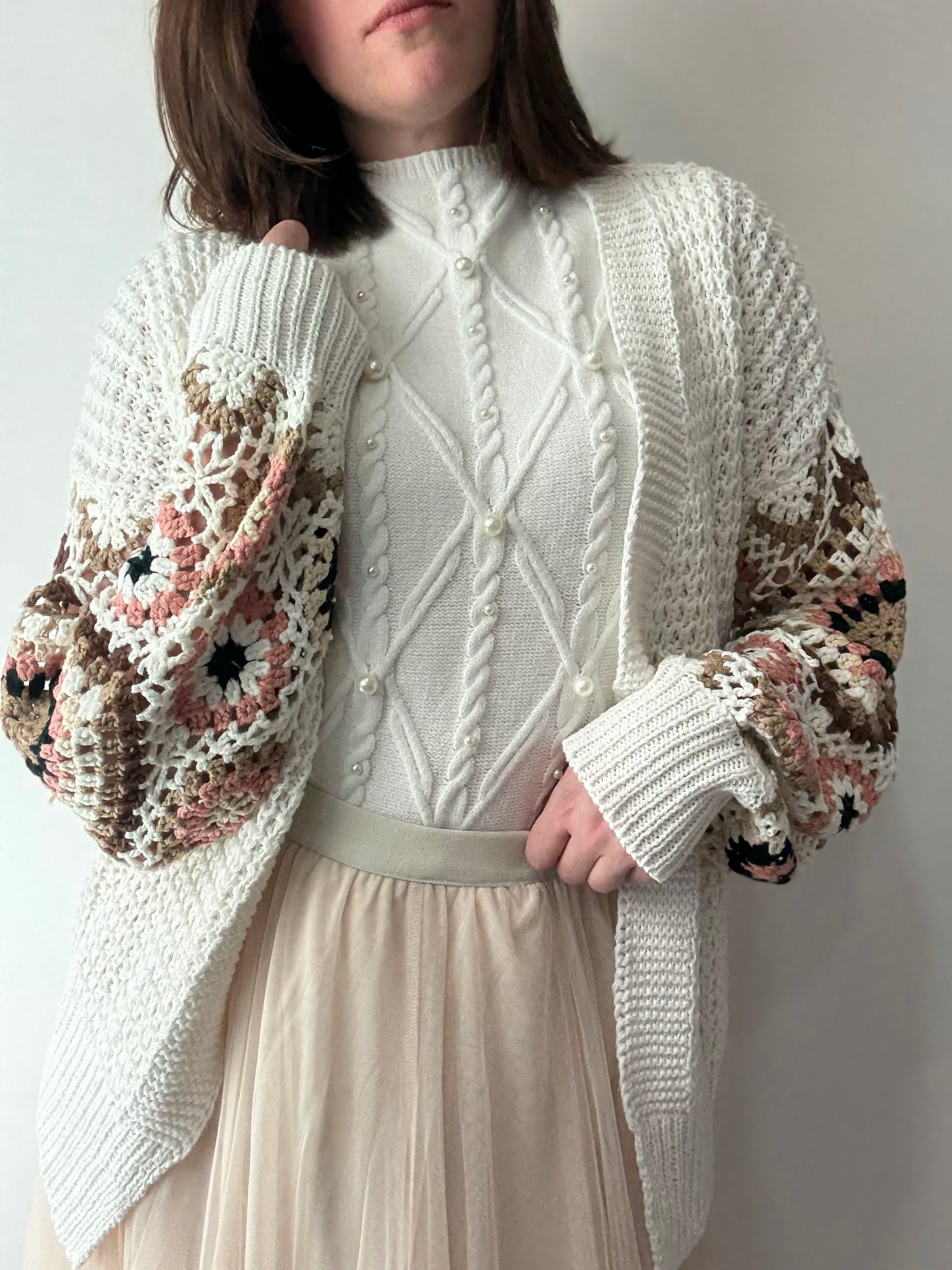 Person wearing a white knitted cardigan with floral patterns on a plain background