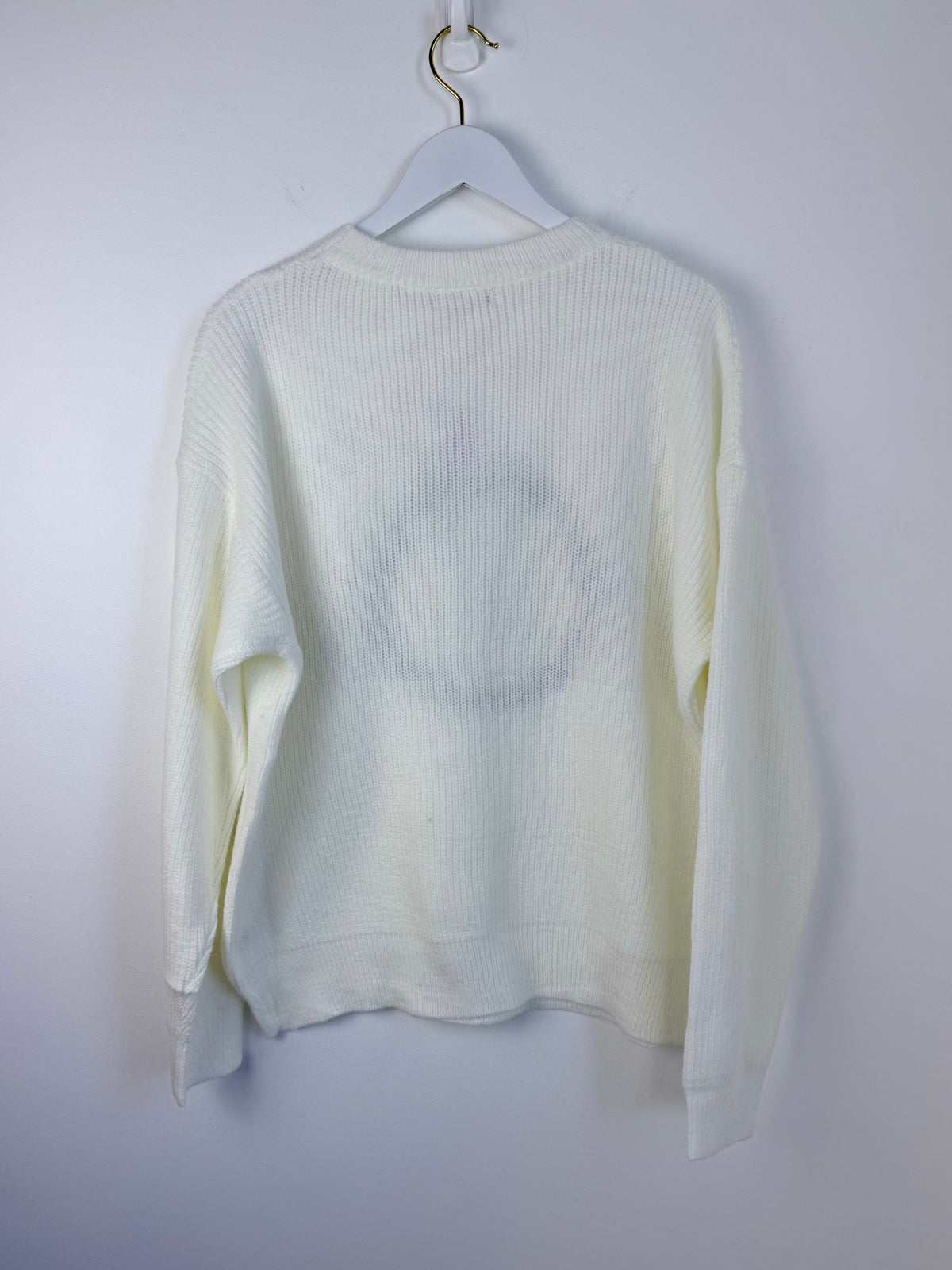 A cream-colored ribbed knit sweater hanging on a white hanger against a plain light gray wall.
