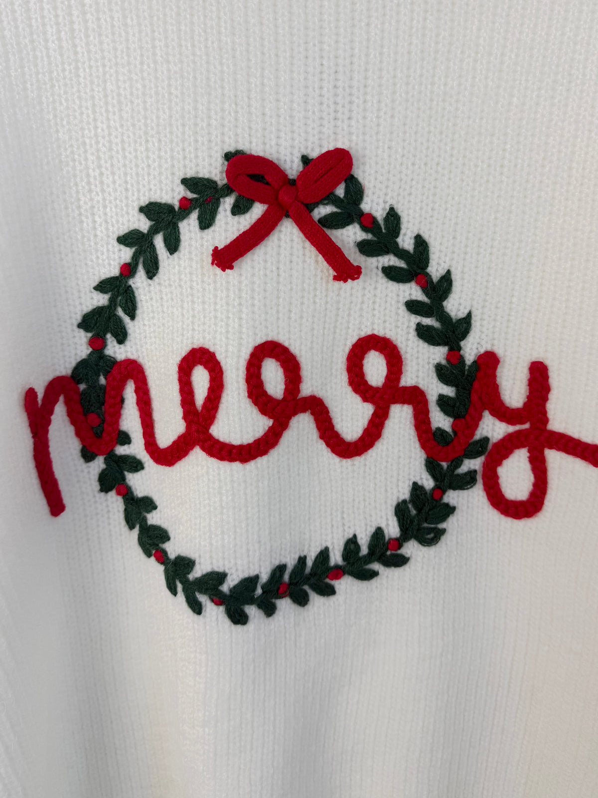 A close-up of a white knit fabric featuring a festive embroidered design with the word 'merry' in red script inside a green wreath with a red bow.