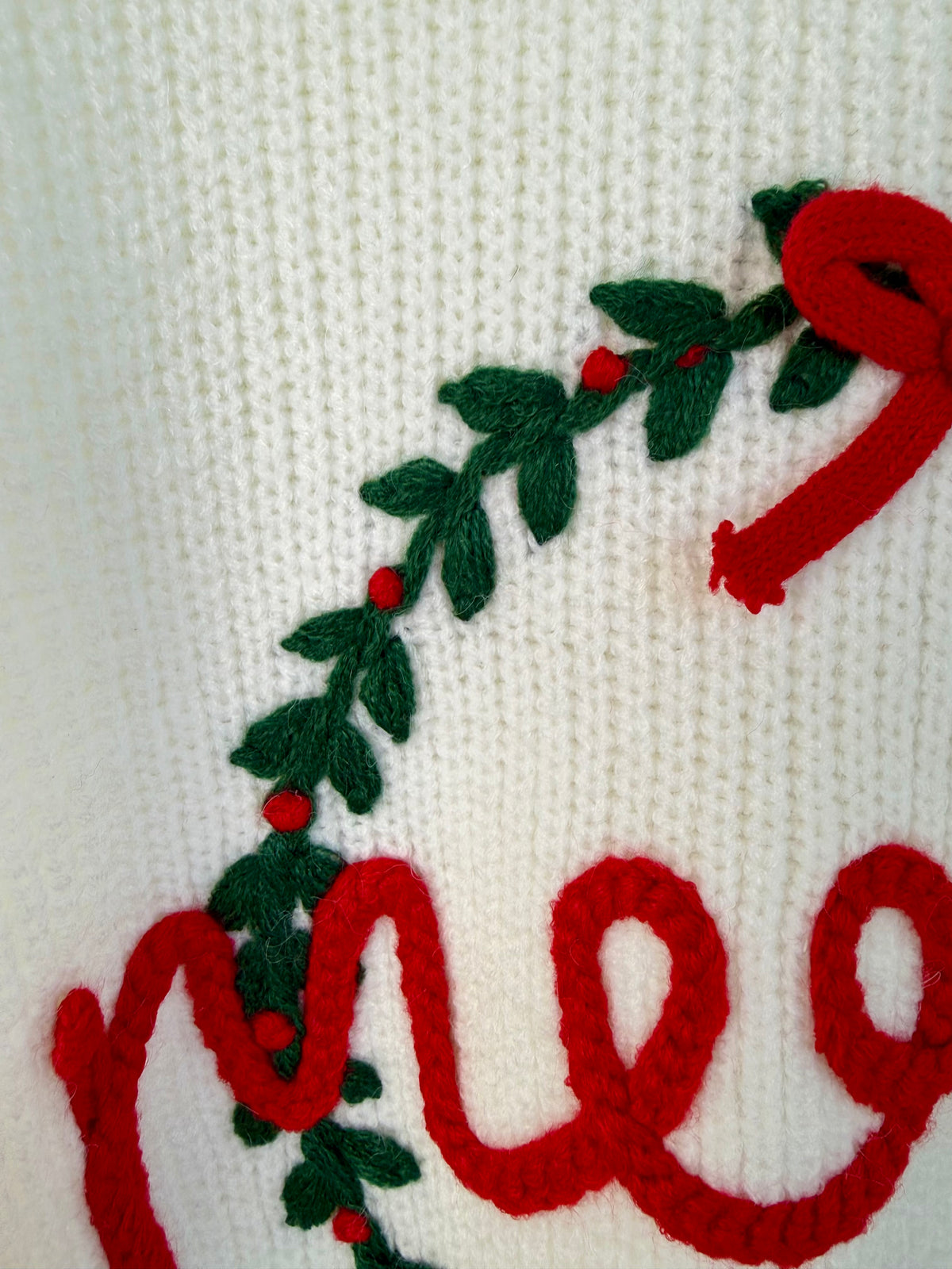 Close-up of a white knit fabric with a red and green embroidered holiday garland and partial red lettering.