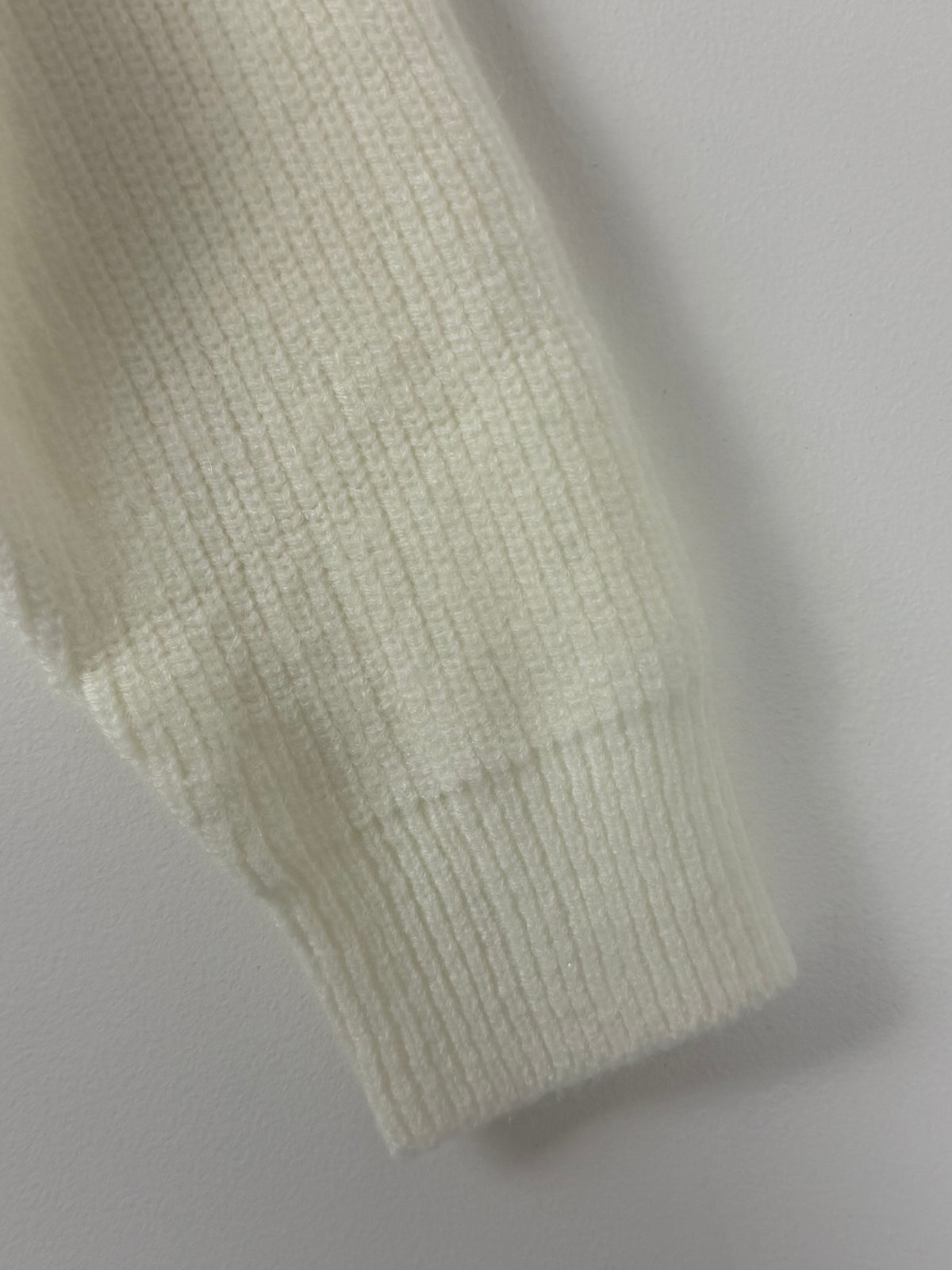 Close-up of a ribbed knit sleeve in a warm off-white or cream color, showing the texture and cuff detail against a neutral light gray background.