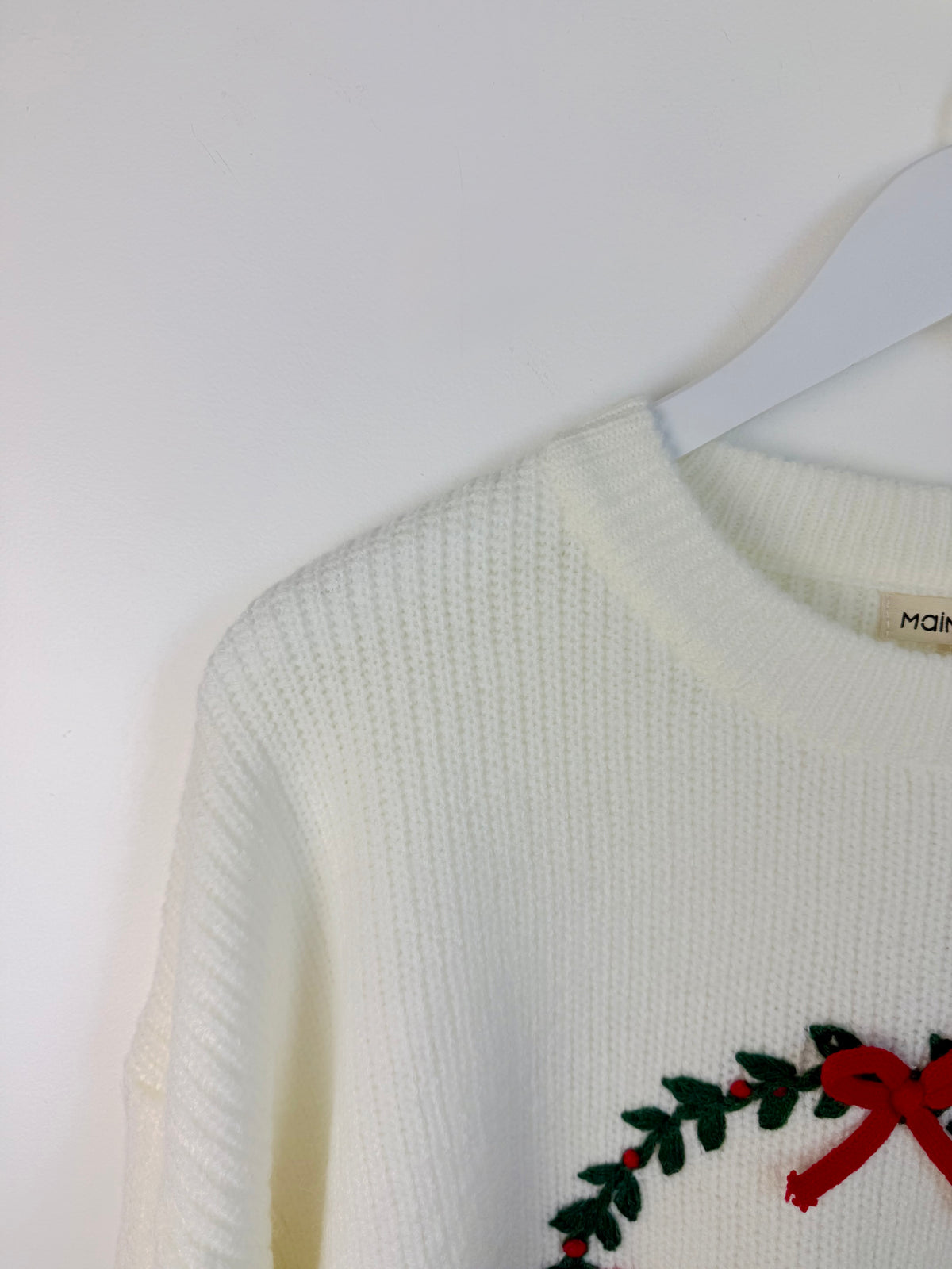 Close-up of a cream-colored ribbed knit sweater hanging on a white hanger against a white wall, featuring a green holly wreath and red bow detail on the lower right.