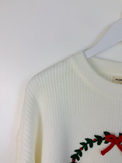 Close-up of a cream-colored ribbed knit sweater hanging on a white hanger against a white wall, featuring a green holly wreath and red bow detail on the lower right.