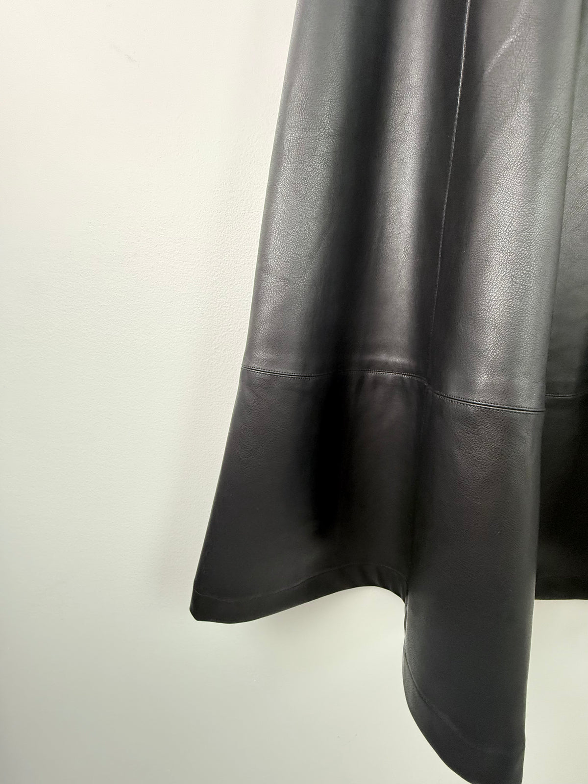 A close-up of a black leather or faux-leather garment, showing the draped fabric and a horizontal seam detail against a plain off-white background.