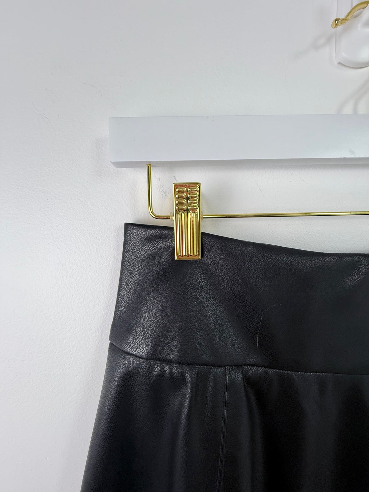 A close-up of a black faux-leather skirt or pants waistband hanging from a gold metal clothes hanger against a white wall.
