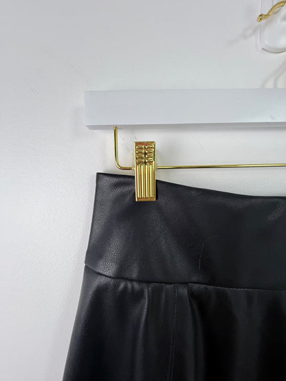 A close-up of a black faux-leather skirt or pants waistband hanging from a gold metal clothes hanger against a white wall.