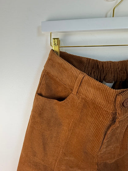 Close-up of brown corduroy pants hanging on a white hanger with a gold clip against a white wall.