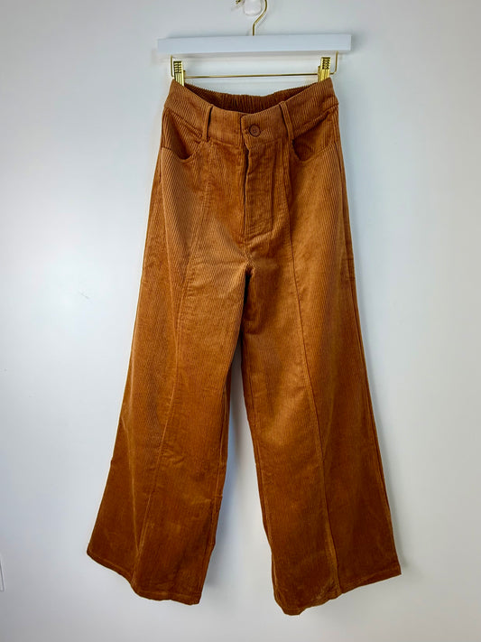A pair of wide-leg brown corduroy pants hanging from a white hanger with gold clips against a plain white wall.