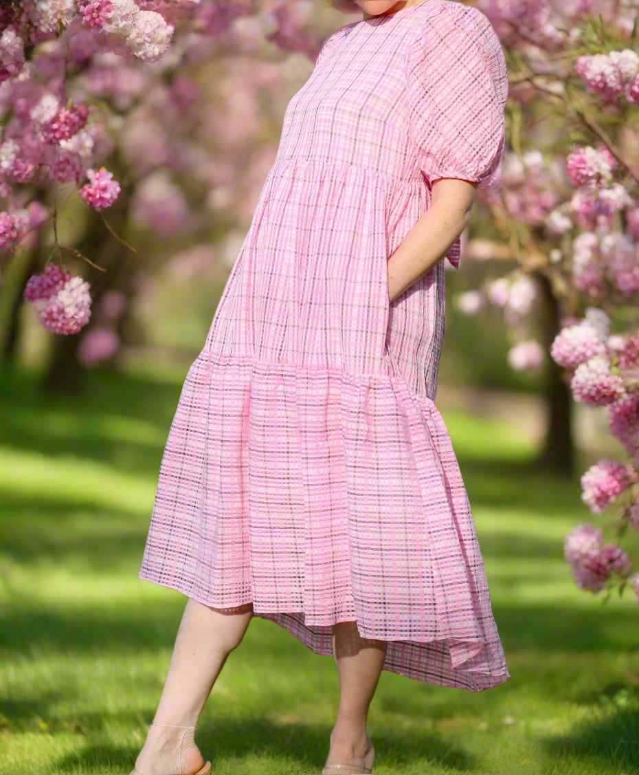 A person standing on grass wearing a pink plaid high low dress with plush puff sleeves and a bow at the back of the neck.