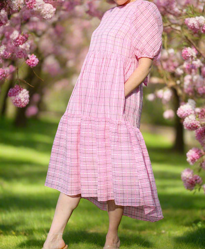 A person standing on grass wearing a pink plaid high low dress with plush puff sleeves and a bow at the back of the neck.