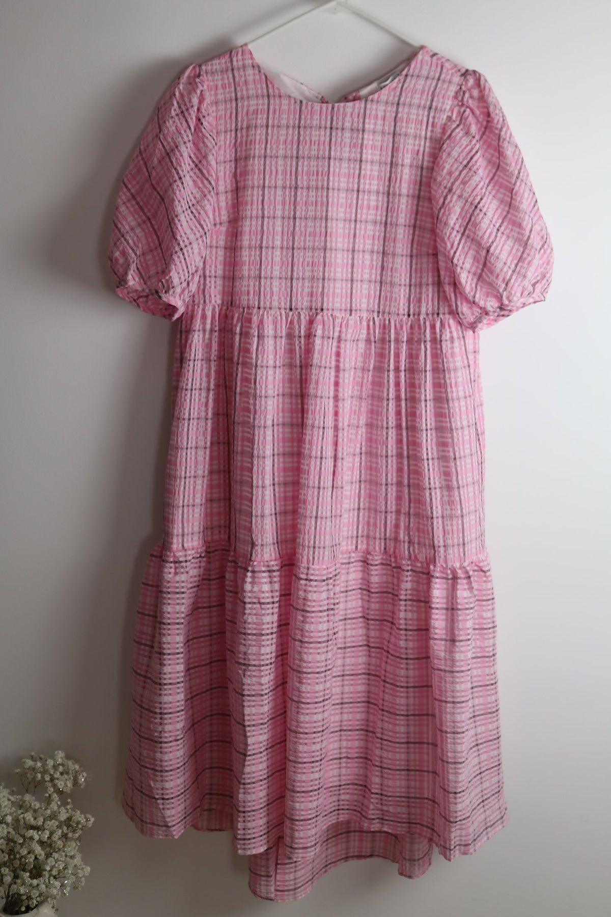 A pink and white plaid tiered dress with short puff sleeves hanging on a white hanger against a plain light gray wall.